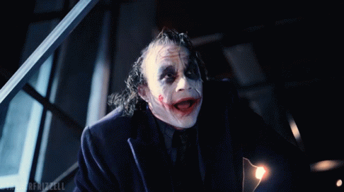Man in white clown face paint and a dark suit grinning and leaning forward in a dim room, in a reaction GIF